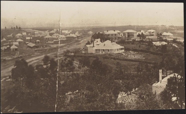 A faded yellow black and white photo shows an early scene of Auckland with single story dwellings and tents scattered along a dirt road along with one or two two-story buildings.