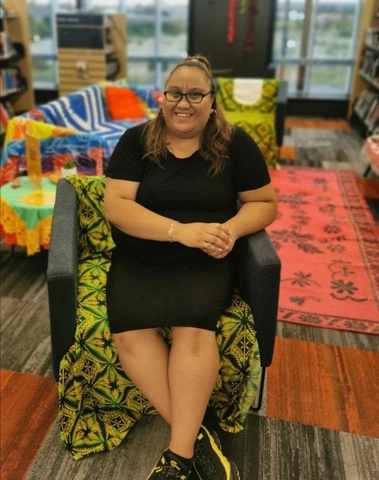 A woman seated in a chair in what appears to be a library setting is smiling with her hands folded in her lap.