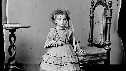 Victorian black and white photo of a young girl standing by a chair. Her hand is in front of her stomach.