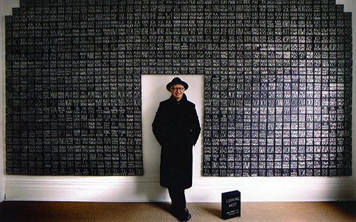 A man wearing a black hat and coat standing in front of an artwork.