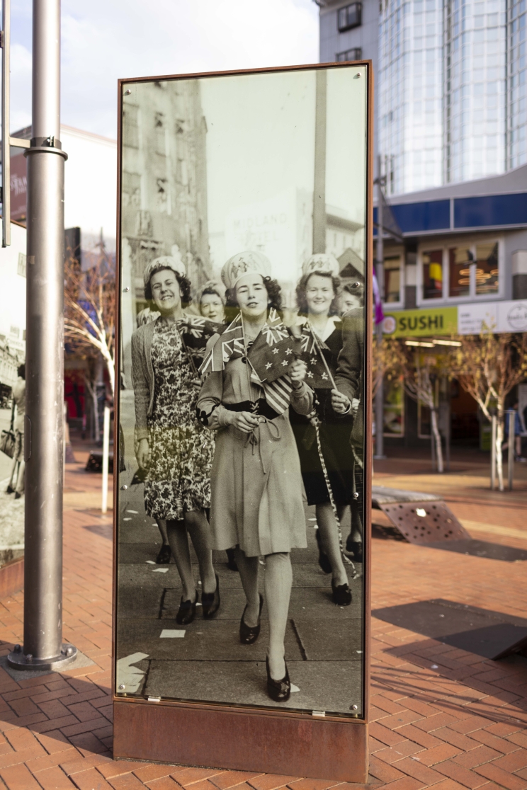 A street scene showing a large light box containing a black and white image of a group of women celebrating and holding small NZ flags.