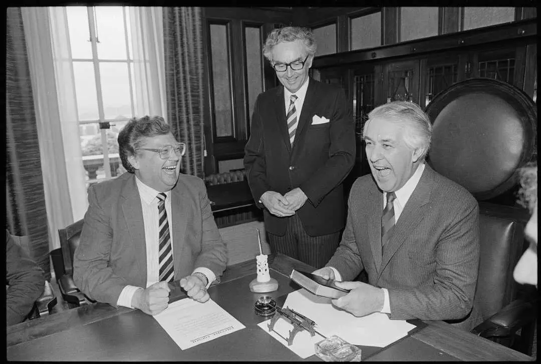 A black and white photo showing an interior office scene with Prime Minister David Lange presenting a bible, pen and pen holder to Governor General Sir Paul Reeves. Also present is Cabinet secretary and clerk of the Executive Council Patrick Millen.