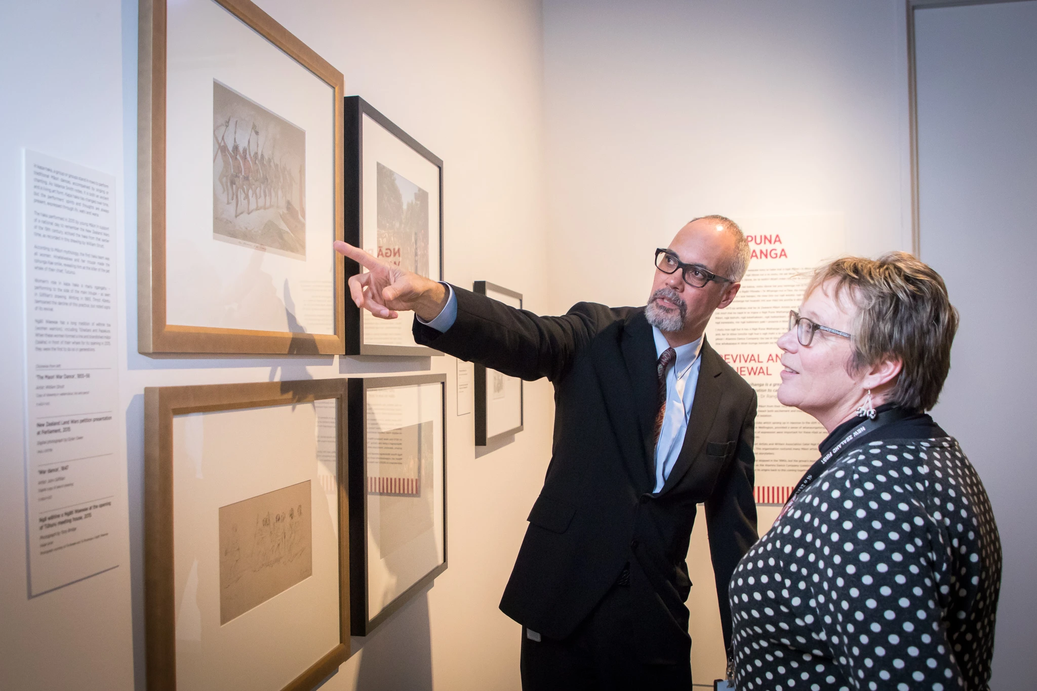 Two people looking at pictures on the wall at the Pūkana exhibition.