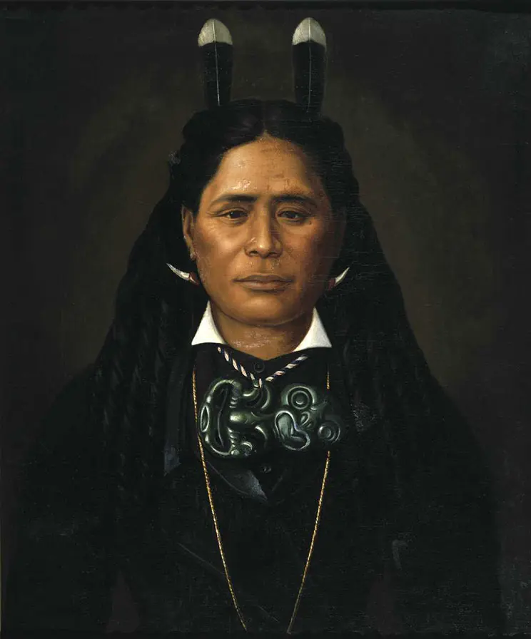 A striking portrait of a Māori woman. She Is wearing dark tailored clothing, has two huia feathers in her hair and a large tiki necklace. She is looking straight at us and has a Mona Lisaish smile.