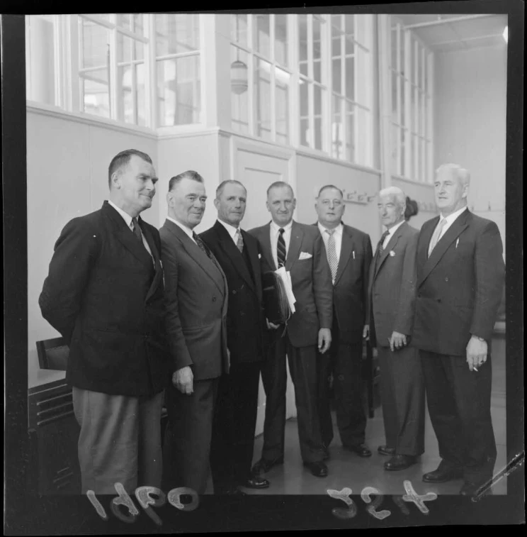 A black and white photo of a group of men in suits standing in a room with a high-ceiling.