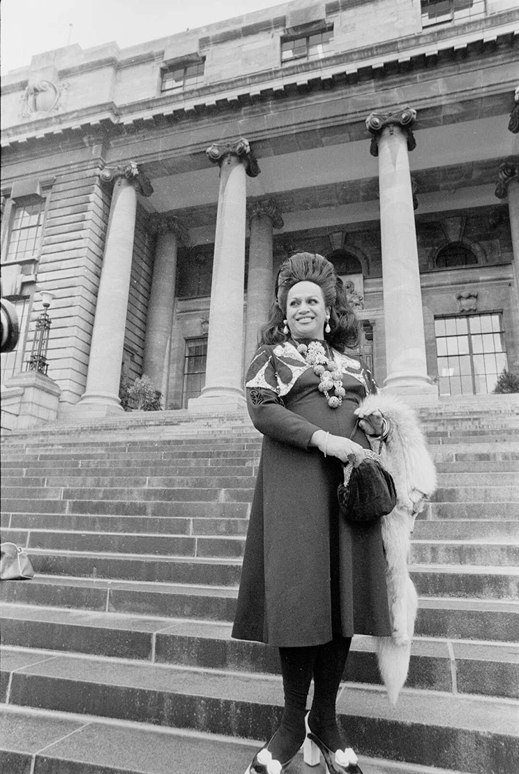 Carmen Rupe standing on the steps of an Edwardian neo classical style building.