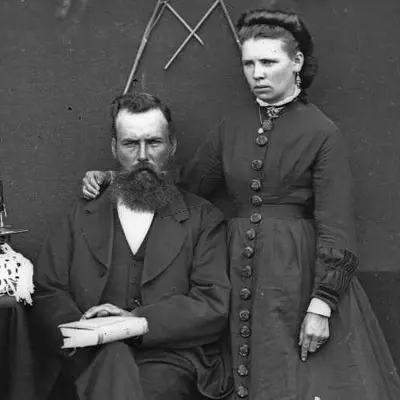 A man and woman wearing 1800s clothing. The woman has her arm around the man. 