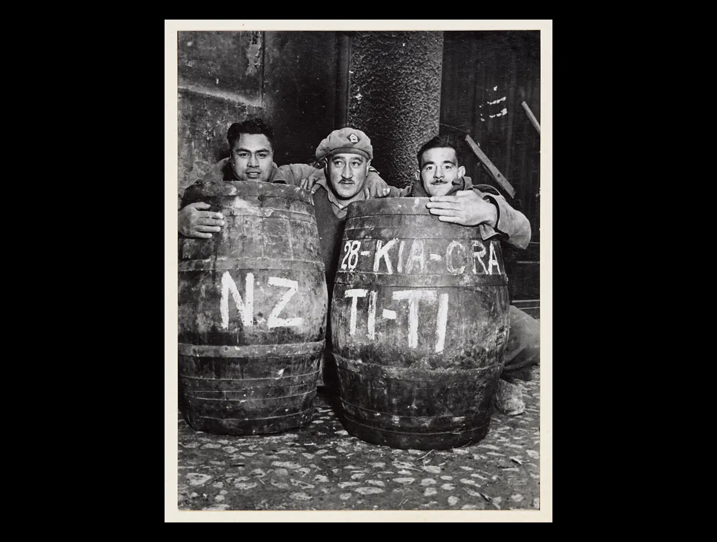 3 soldiers from the 28th (Māori) Battalion peering out from behind 2 barrels of tītī (muttonbirds) sent from Aotearoa NZ.