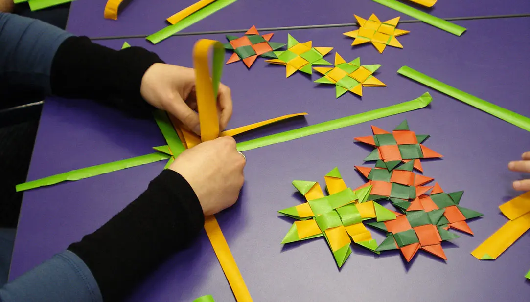 Photo of a person weaving a Matariki star with paper on a table. On the table, it shows other stars already completed.
