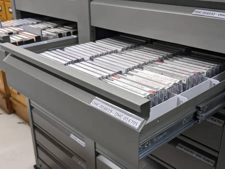 A row of cassettes are neatly stored in grey archival drawers with labels on them. 