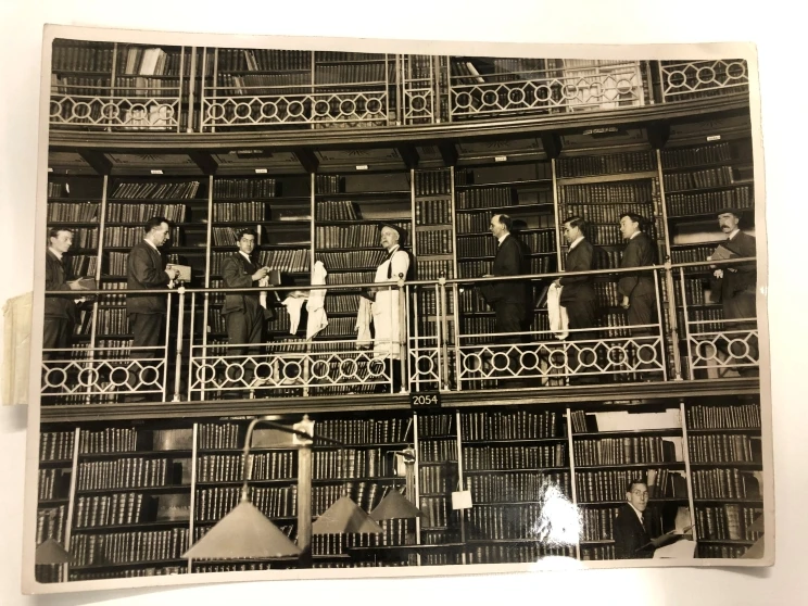 Black and white photo showing a library with shelves lined with books while half a dozen men stand with dust clothes dusting the books and posing for the camera.