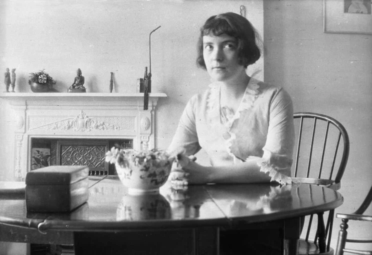 Black and white photo of a woman seated at a kitchen table with a bowl of flowers  in front of her and a fireplace in the background.