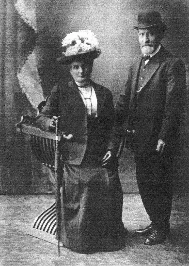 A formal black and white portrait photograph of a couple, with the woman seated and the man standing beside her.