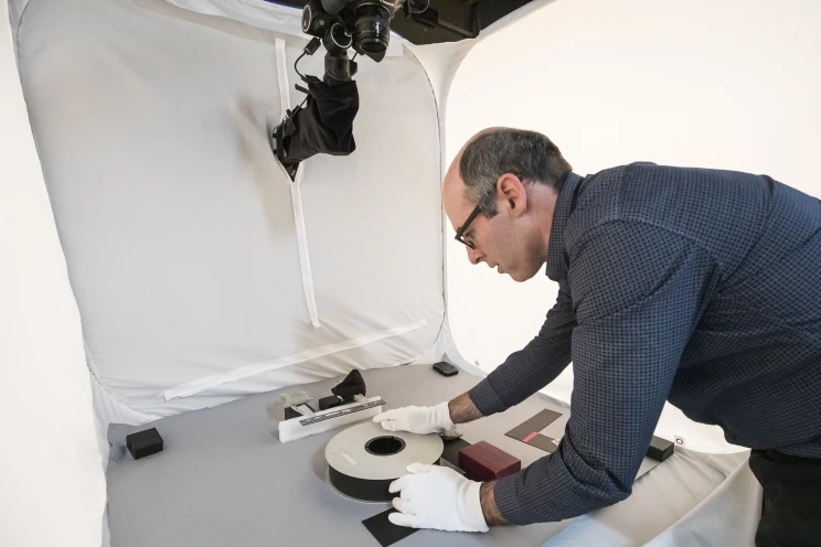 A reel of magnetic tape is displayed on grey hardboard being positioned by photographer wearing white gloves.