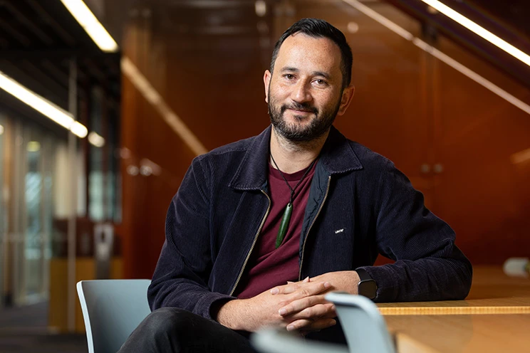 A smiling Māori man wearing a pounamu seated casually at a table.