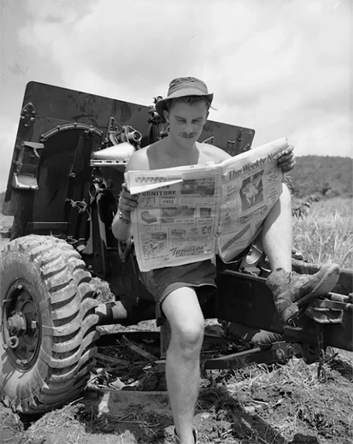 Black and white photo of a man wearing a hat, shorts and muddy boots, sitting on a field gun, reading a newspaper.