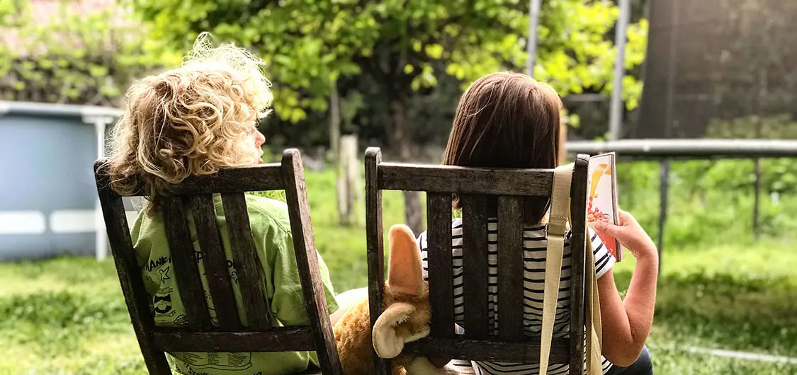 2 tamariki (children) sitting on chairs in a yard. One is reading a book.