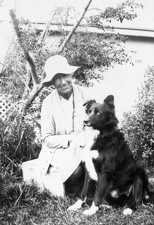 Black and white photo of a woman wearing a floppy hat crouched down next to a dog in a garden.
