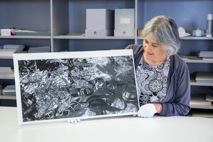 A woman wearing white gloves holds a large black and white photographic print.