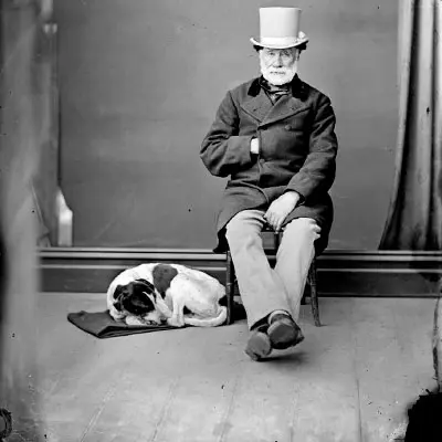 Black and white photo of a seated man wearing a top hat. His dog is curled up next to him. 