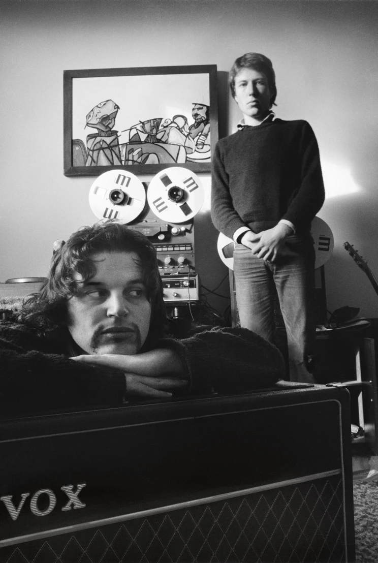 A black and white photo of Chris and Alec in their recording studio, Chris is resting his head on a large guitar amp and Alec is standing next to a reel-to-reel tape recorder.