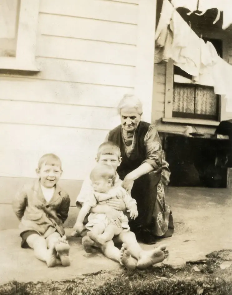 A woman and three young children out front of a wooden house with laundry hanging on the bannister in the background. 