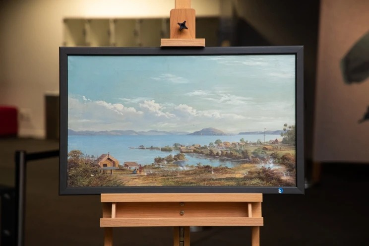 An oil painting on an easel showing a summery-looking scene of a small village next to a lake.