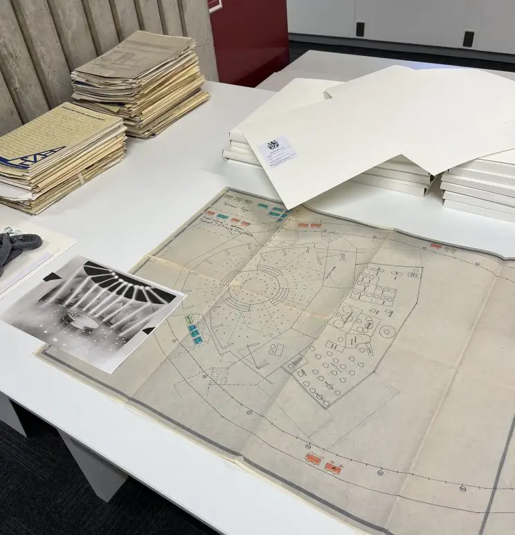 On a table are various piles of papers and folders along with a stage plan and photos of what appears to be the same stage setup.