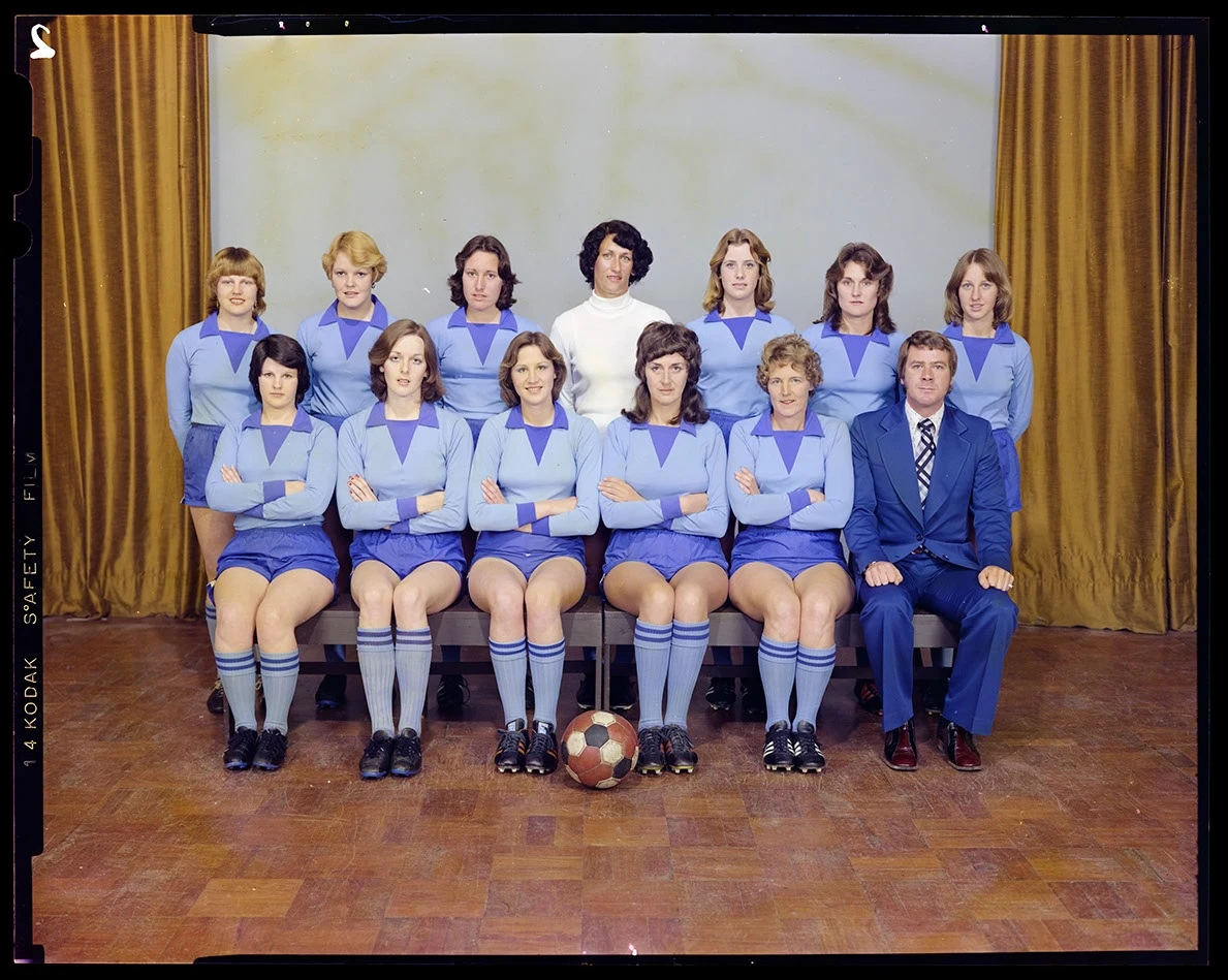 A colour studio portrait of a women's soccer team with two rows of players in blue uniforms including the captain in a white jersey and a male coach seated at bottom right. 