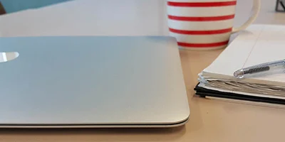 A desk with a pen, notebook, cup, and laptop on top.