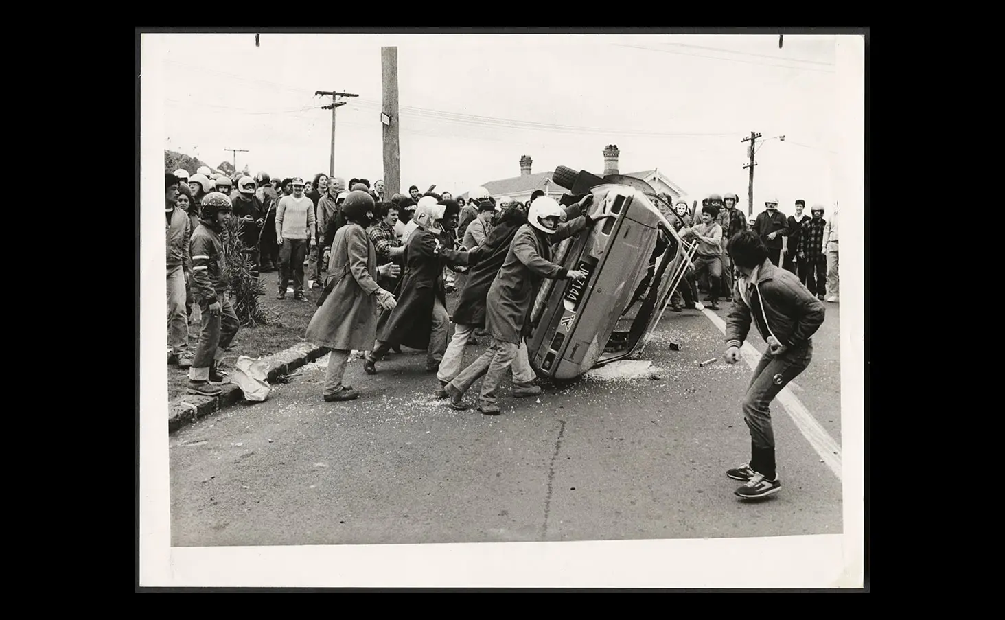 Black and white photo of 1981 anti-Springbok tour supporters flipping over a car on an Auckland street. Some are wearing motorcycle helmets for protection.