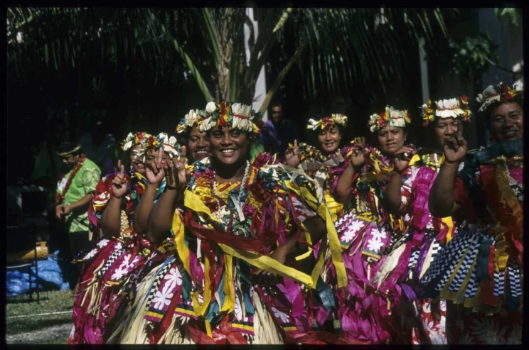 Women lined up in rows and wearing traditional dress with bright colours and flowers appear to be smiling and dancing.