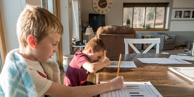 2 boys sitting at a dining table doing schoolwork. The boy in the foreground has a blanket wrapped around him.