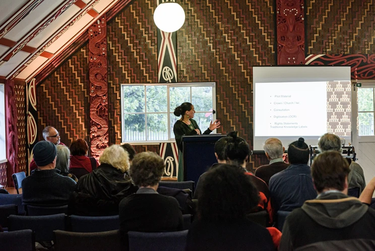 A woman gesturing at a screen while presenting to a group of seated people in a marae.