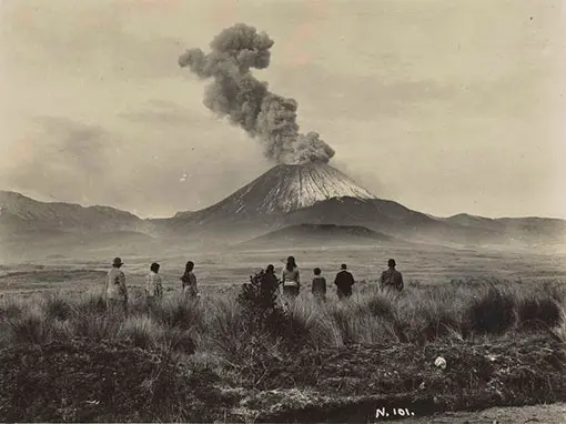 People watching a conical shaped mountain with large plumes of steam coming from its centre. 