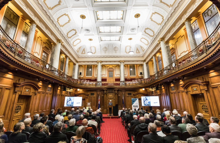 View from the back of the Grand Hall, full with people seated in rows with an aisle down the middle showing red carpeting.