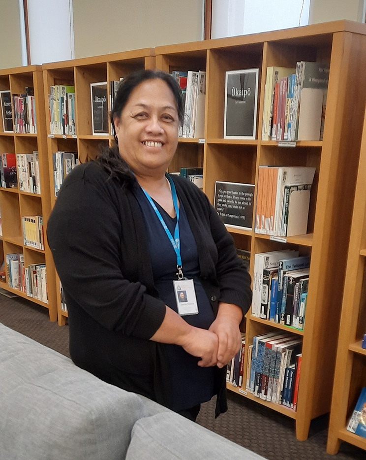 Māori woman standing in from of bookshelves. 