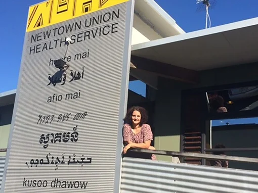 A woman leaning against a sign with the text 'Newtown Union Health Service' and 'welcome' in 7 different languages.