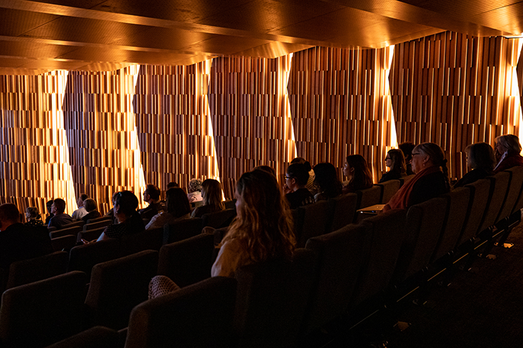 People sitting in a dimly lit auditorium with beautiful decorative wood panels highlighted. 