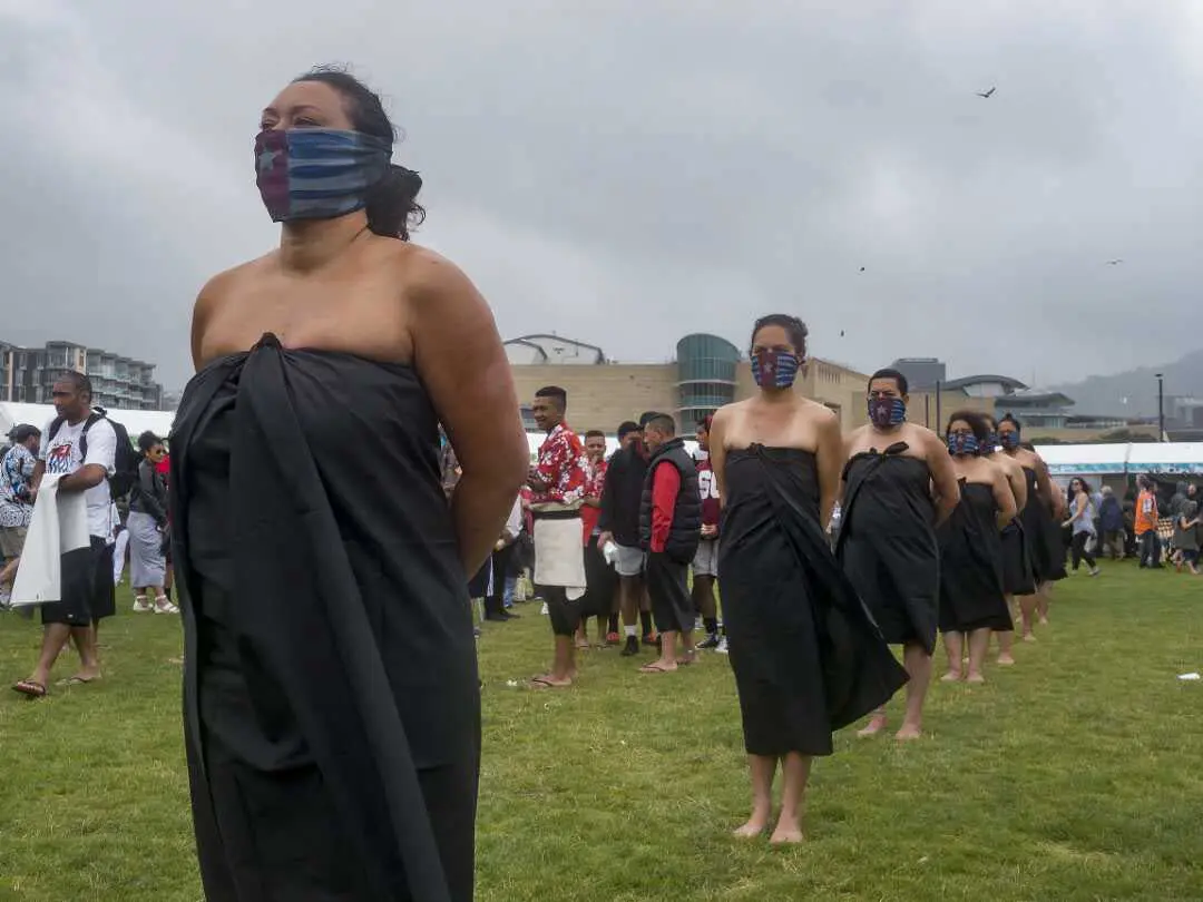 Women in a line wearing black wraps around their torsos, shoulders are bare. They have flags covering the lower part of their faces. Their hands are behind their backs.