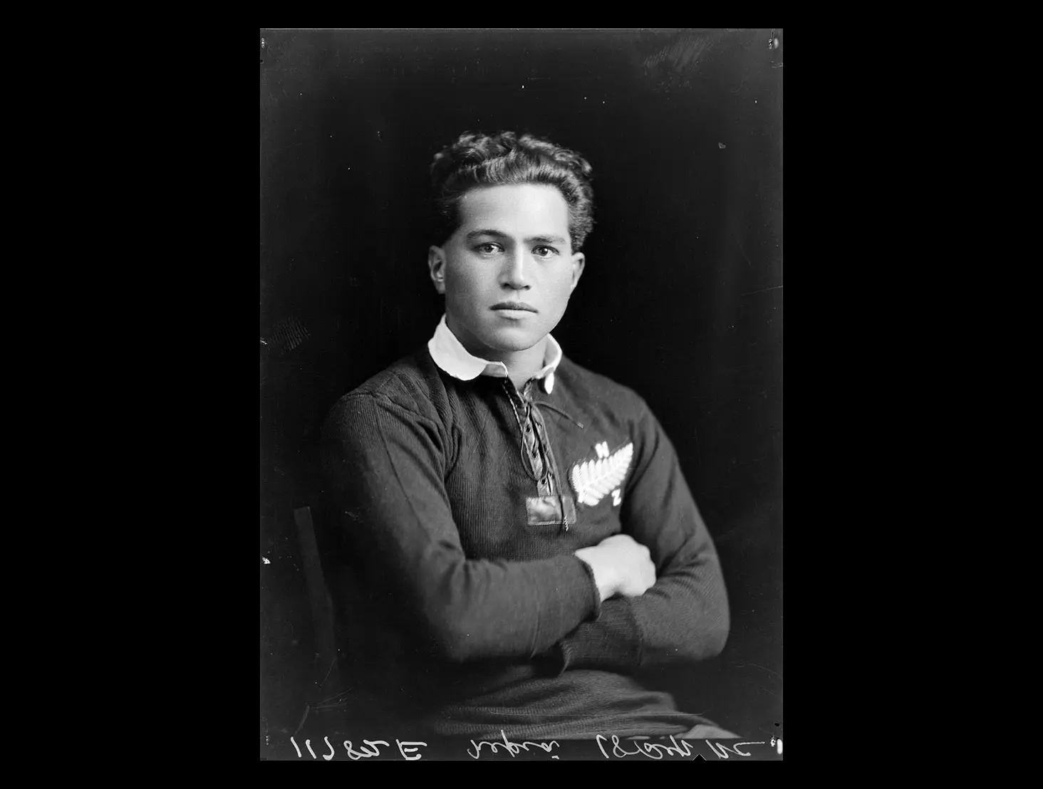 Studio back-and-white photo of George Nēpia, wearing an All Blacks uniform, with arms crossed looking at the camera.