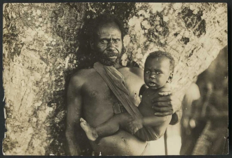 A man stands against a large tree whilst holding a baby on his hip which is also supported with a cloth sling.