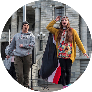 Two Māori people standing in front of Parliament. One has a Tino Rangatiratanga flag on as a cape.