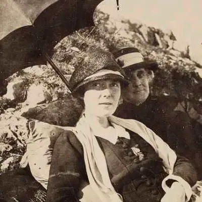 Two women in early 1900s clothing at the beach sitting under an umbrella.