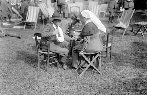 A nurse and two wounded soldiers seated in a group on wooden chairs on the grass.