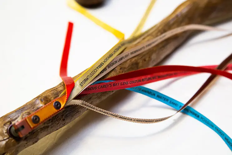A polished piece of thin wood with thin ribbons on it that are coloured and include writing connected to it.