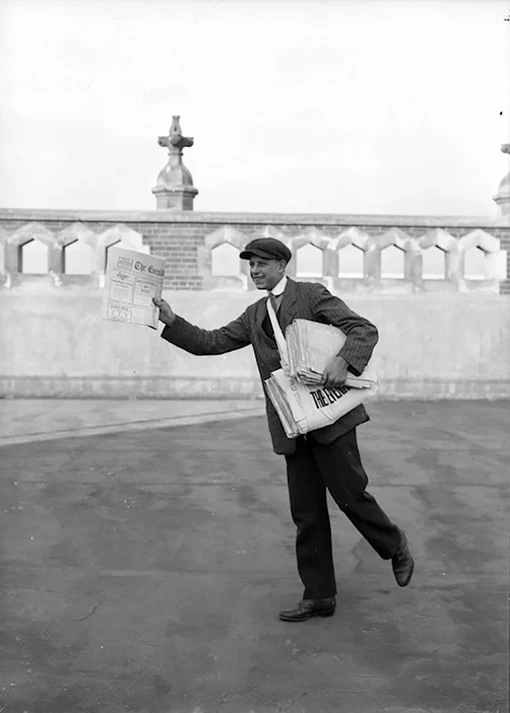 Black and white photo of a young man wearing a suit and peaked cap, holding a bundle of newspapers for sale.