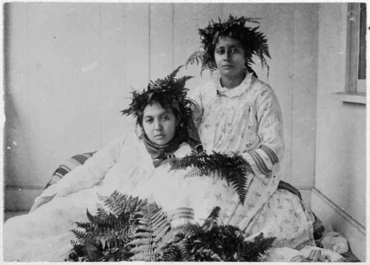 A black and white portrait of two women sitting on the ground with a headdresses made of ferns.