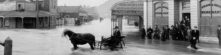 Flooded town street, a horse and cart stands in the floodwaters, and people are grouped outside a building. 