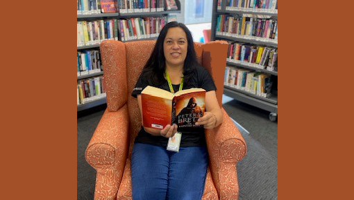 Woman sitting in a chair reading a book. There are bookshelves behind her. 